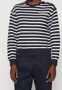 Sweatshirt rayé bleu marine et blanc en coton, avec un col rond côtelé, des boutons décoratifs sur une épaule et un logo brodé.