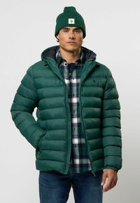 Groene gewatteerde jas met een gequilt patroon, rits aan de voorkant en capuchon. Draag met een blauw-groene geruite shirt, gecombineerd met blauwe jeans en een groene beanie.
