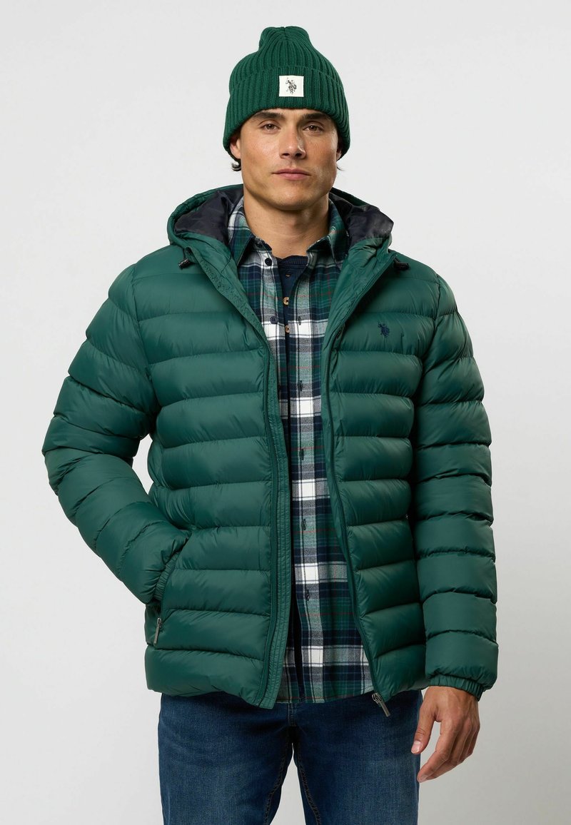 Groene gewatteerde jas met een gequilt patroon, rits aan de voorkant en capuchon. Draag met een blauw-groene geruite shirt, gecombineerd met blauwe jeans en een groene beanie.