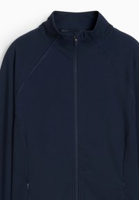 Sinine lukuga jakk, mis on valmistatud siledast kangast. Omab kõrget kaelust ja raglansleeve'i. Sisaldab kahte lukuga küljetaskut.