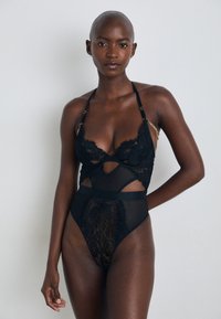 Juoda nėrinių bodysuit su iškarpų dizainu, reguliuojamomis petnešėlėmis ir auksiniais akcentais. Permatoma medžiaga su gėlių raštais ir aukštu liemeniu.