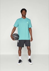 Hombre con camiseta verde azulado, pantalones cortos negros y deportivas blancas sosteniendo un balón de baloncesto negro, de pie contra un fondo claro y liso.
