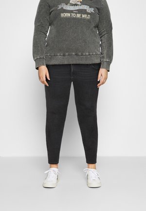 Person iført falmet sort sweatshirt med ørn og teksten 'Born to Be Wild', sorte jeans og hvide sneakers, stående på gråt gulv.