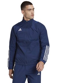 Marineblaue Sportjacke mit hohem Kragen, durchgehendem Reißverschluss und drei weißen Streifen an den Ärmeln. Mit einem Adidas-Logo auf der Brust.