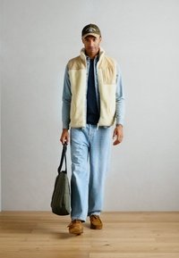 Beige fleece vest over a blue button-up shirt, light blue jeans, brown shoes, and a green bag. The vest has large pockets and a textured finish.