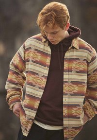 Jonge man met rood haar in een gestippeld hemd over een bordeauxrode hoodie, die naar beneden kijkt met zijn handen in zijn zakken.