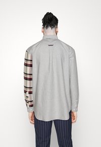 Langarmhemd mit einem gestreiften grauen Körper und einem kontrastierenden Karomuster am linken Ärmel. Verfügt über eine geknöpfte Manschette und eine Rückenfalte.