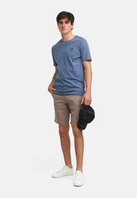 T-shirt en coton bleu avec un col rond, shorts beiges et baskets blanches ; tenant une casquette à carreaux sombres, debout devant un fond uni.
