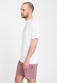 Homme avec une barbe courte portant un t-shirt blanc et un short rose se tenant de profil devant un fond blanc uni.