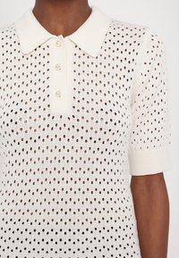 Gros plan d’un polo en maille blanche à manches courtes, avec de petits trous, un col plié et trois boutons en nacre.