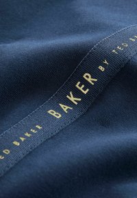 Mornarski materijal s glatkom teksturom, s ušivenom etiketom na kojoj piše "BAKER by TED BAKER" zlatnim slovima uz rub.
