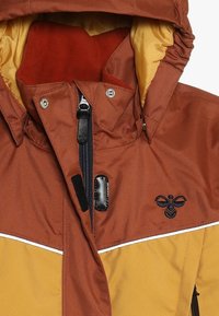 Giacca isolante marrone e gialla con cappuccio, chiusura a zip e bottoni a pressione. Presenta dettagli neri e logo sul petto.