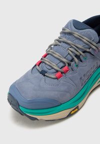 Blå semsket sneakers med flerfargede snørebånd, teksturert overdel, fremtredende mellomsole-lag i grønn og brune nyanser, samt Vibram-merke på sålen.
