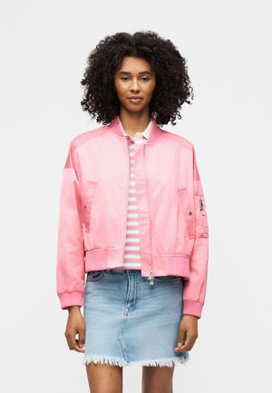 Tânără cu păr creț purtând o jachetă bomber roz, o bluză cu dungi și o fustă din denim cu tiv desfăcut, stând pe un fundal alb.