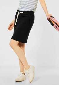 Personne portant une jupe noire à mi-mollet, des baskets blanches et une chemise rayée, tenant une écharpe colorée dans une main.