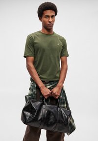 Μαύρη υφαντή τσάντα duffel με λείες δερμάτινες λεπτομέρειες, με στρογγυλεμένο σχήμα και ανθεκτικές λαβές. Συνδυάζεται με πράσινο ελιάς t-shirt.