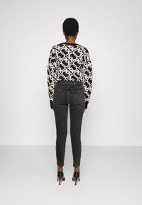 Person med kort hår som bär en svart och vit mönstrad tröja, mörkgrå tajta jeans och svarta högklackade skor, stående med ryggen vänd.