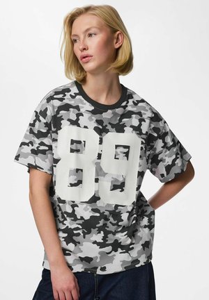 Lyshåret kvinde iført en grå sort og hvid camouflage t-shirt med stort hvidt nummer 89, poserer med hånden på hoften og kigger mod venstre.
