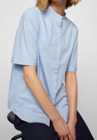 Hellblaues Kurzarmhemd mit Mandarin-Kragen, aus glattem Stoff gefertigt. Verfügt über eine gerade Passform und dezent ausgearbeitete Seitennahtdetails.