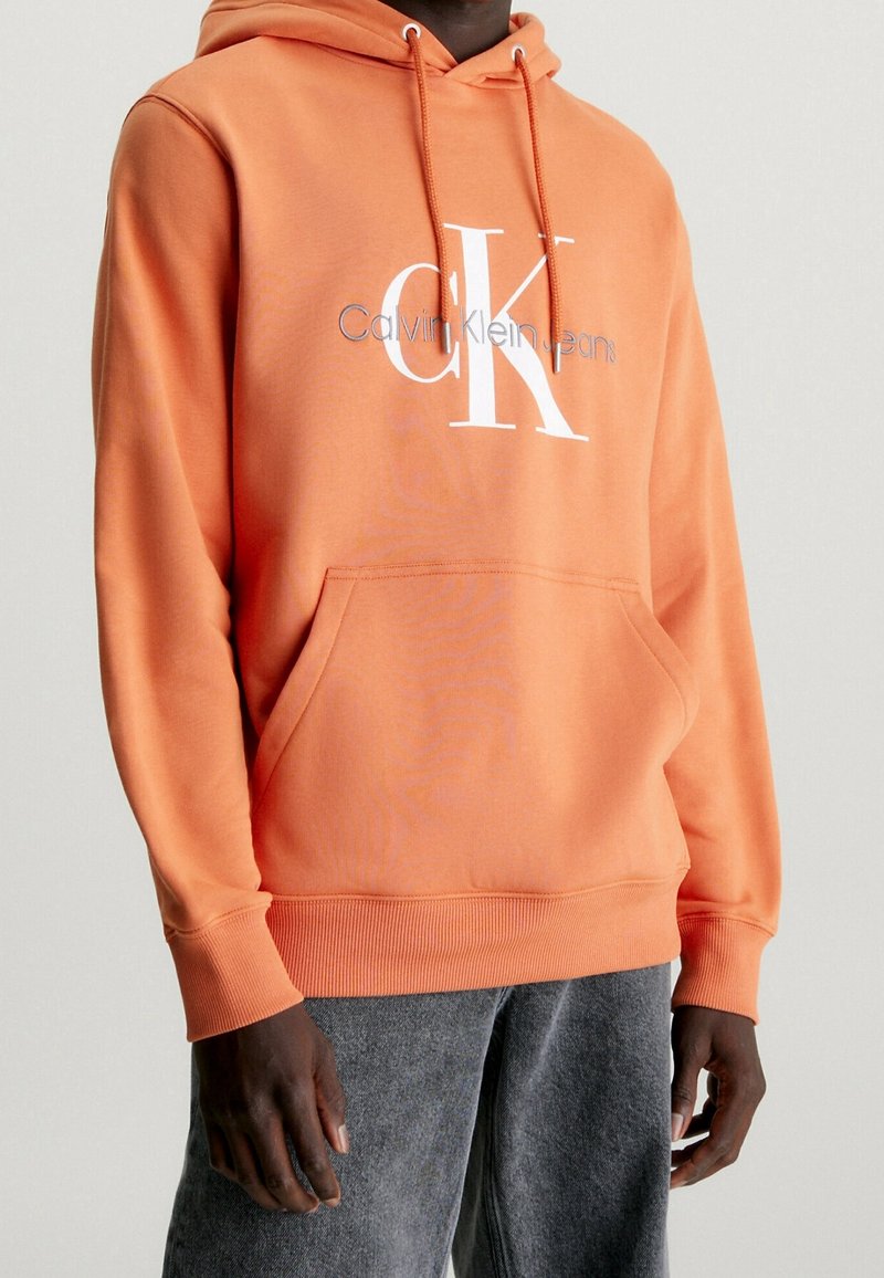 Orange hoodie made of cotton blend, featuring a front kangaroo pocket and a large white logo with “Calvin Klein Jeans” text.