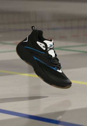 Baskets Reebok noires et bleues avec languette blanche, flottant au-dessus d’un sol de gymnase marqué de lignes de terrain de sport en bleu, jaune et vert.