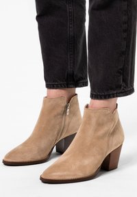 Beige Wildleder-Ankle-Boots mit spitzer Zehenform, Reißverschluss an der Seite und einem gestapelten Holzabsatz, kombiniert mit schwarzen, cropped Jeans.