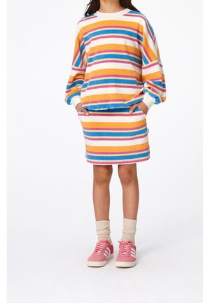 Enfant portant un pull à rayures colorées et une jupe assortie, les mains dans les poches, avec des chaussettes beiges et des baskets Adidas roses.