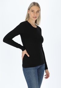 Maglietta nera a maniche lunghe con scollo rotondo, design aderente e texture liscia, abbinata a jeans blu, su uno sfondo bianco semplice.