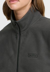 Hallhall fliisist zip-up jakk, millel on kõrge krae, sujuva tekstuuriga ja silmapaistev "OXMO" logo rinnal.