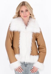 Blonde Frau trägt eine beige Jacke mit flauschigem weißen Kragen und Manschetten, kombiniert mit hellblauen Jeans, posiert mit den Händen nahe der Taschen.