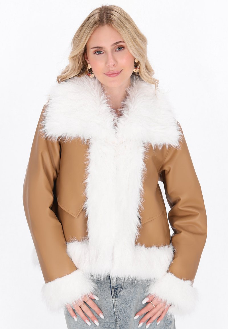 Blonde Frau trägt eine beige Jacke mit flauschigem weißen Kragen und Manschetten, kombiniert mit hellblauen Jeans, posiert mit den Händen nahe der Taschen.