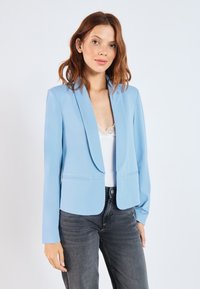 Blazer ajusté bleu clair avec un col cranté, une fermeture à un bouton à l'avant et deux poches avant. Porté sur un débardeur en dentelle blanche et un jean foncé.