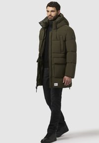 Oliven grøn puffjakke med høj krave, sidelommer og lynlås foran. Kombineret med sorte jeans og mørke sneakers.