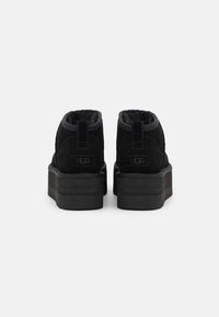 Botines de ante negro con un interior acolchado, suela gruesa de plataforma negra y un logo de UGG bordado en el talón. Diseño simple y moderno.