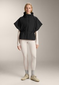 Zwarte capuchonjas met ritssluiting aan de voorkant, een geplooide taille en brede mouwen, gecombineerd met lichtbeige leggings en grijze enkelsokken.