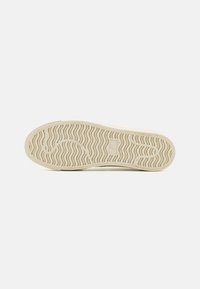 Semelle d'une basket beige avec un motif ondulé et une zone rectangulaire pour le logo au centre, montrée sur fond blanc.