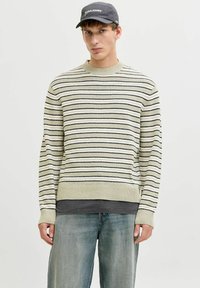 Jeune homme portant un pull à rayures cr ème et noir, un jean bleu délavé, un sous-t-shirt gris foncé et une casquette noire Jack & Jones, debout dans une posture neutre.