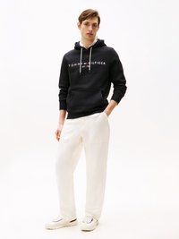 Juodas džemperis su balta "TOMMY HILFIGER" logo, su raišteliais ir priekiu kišenėje. Suderintas su baltais laisvais kelnėmis ir baltais sportbačiais.
