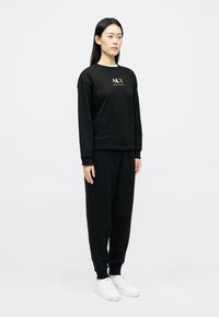 Γυναίκα που στέκεται φορώντας μαύρη μπλούζα Armani Exchange, ταιριαστά μαύρα παντελόνια και λευκά αθλητικά παπούτσια σε απλό ανοιχτό φόντο.