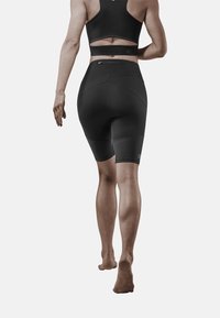 Kobieta idąca boso w czarnym sportowym biustonoszu i rowerowych szortach o podwyższonym stanie z małą kieszonką na zamek z tyłu.