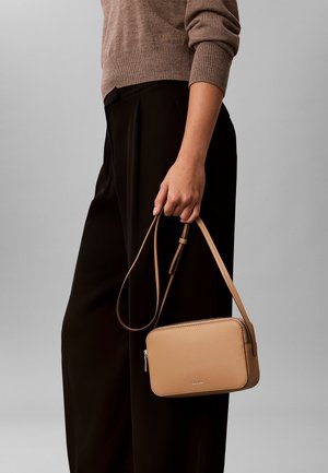 Persona con un suéter marrón y pantalones negros sosteniendo una pequeña bolsa cruzada de cuero beige con cremallera y correa ajustable.