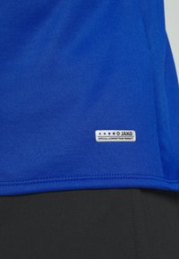 Tissu bleu avec une petite étiquette de marque JAKO indiquant "Official Licensed Team Product" près de l'ourlet au-dessus du tissu noir.
