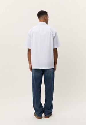 Άνδρας που στέκεται με το βλέμμα στραμμένο προς τα πίσω, φοράει άνετο λευκό πουκάμισο με κοντά μανίκια και διακριτικές ρίγες, μπλε τζιν και καφέ παπούτσια.