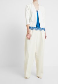 Žena oblečená v bílých širokých kalhotách, bílém krátkém saku a modré tílko s krajkovými detaily, stojící proti jednoduchému bílému pozadí.