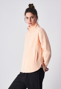 Chaqueta de cremallera en color durazno claro con cuello alto, bolsillos laterales y un corte holgado. Textura suave y detalle de dobladillo redondeado. Combinada con pantalones negros.