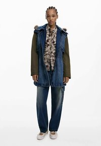 Ilgas džinsinis švarkas su žaliu audiniu viršuje, puoštas kailiu aptraukta gaubtu. Modelis vilki raštuotą marškinius ir laisvus tamsius džinsus su nudilusiais elementais.