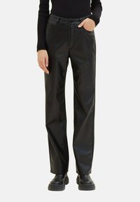 Personne portant un pantalon droit en faux cuir noir, un haut à manches longues noir et des bottes épaisses noires, une main dans la poche, debout sur fond blanc.