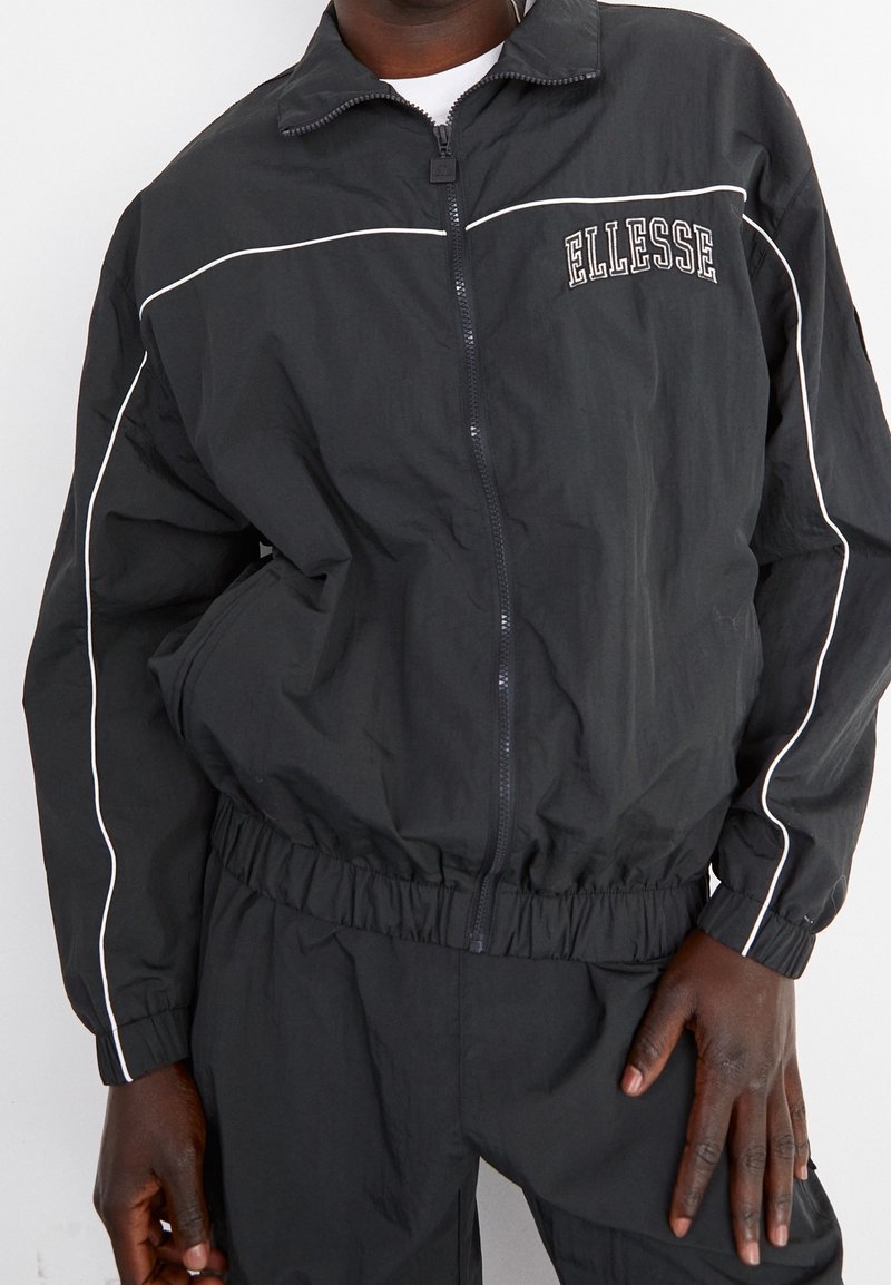 Veste coupe-vent noire avec poignets et taille élastiques, décorée de passepoils blancs et du logo « Ellesse » sur la poitrine. Tissu léger et texturé.
