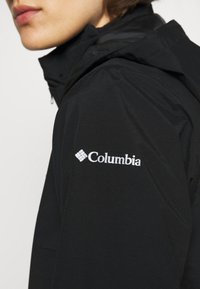 Svart Columbia-jacka med hög krage, dragkedjestängning och broderad logotyp på vänster ärm. Slät, lätt tygstruktur.