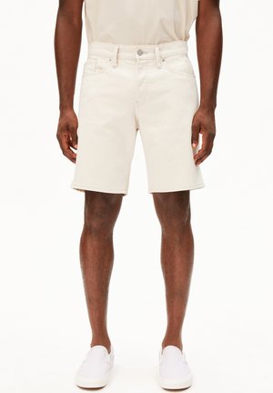 Man die gebroken-witte denimshorts met voorzakken en witte instapschoenen draagt, staand tegen een effen achtergrond.